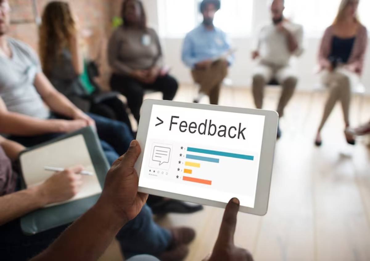 A person holds a tablet displaying a feedback form with ratings, while a group sits in discussion in the background.