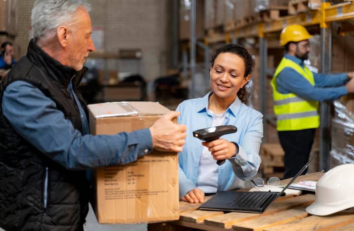 A warehouse scene showing a person checking a box with a scanner while another stands nearby, with a laptop and safety gear on the table.
