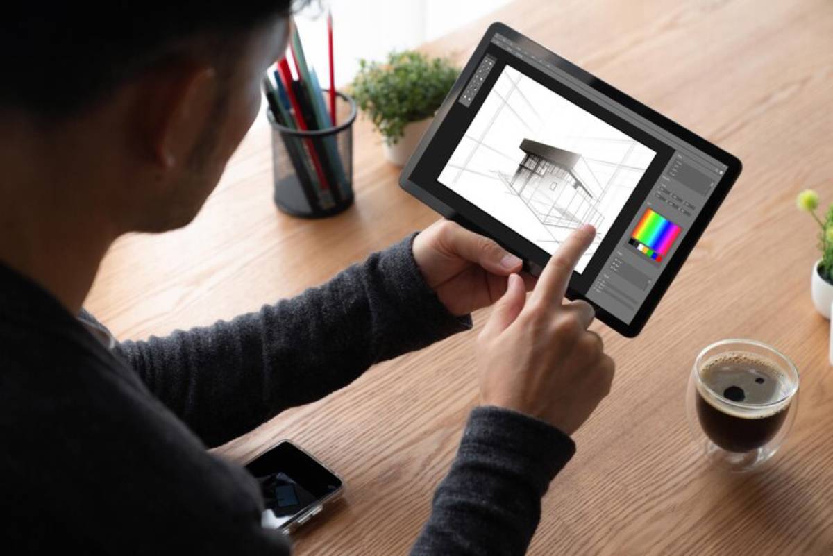A person designs architectural sketches on a tablet, coffee and smartphone on the desk, surrounded by plants and colorful pens.