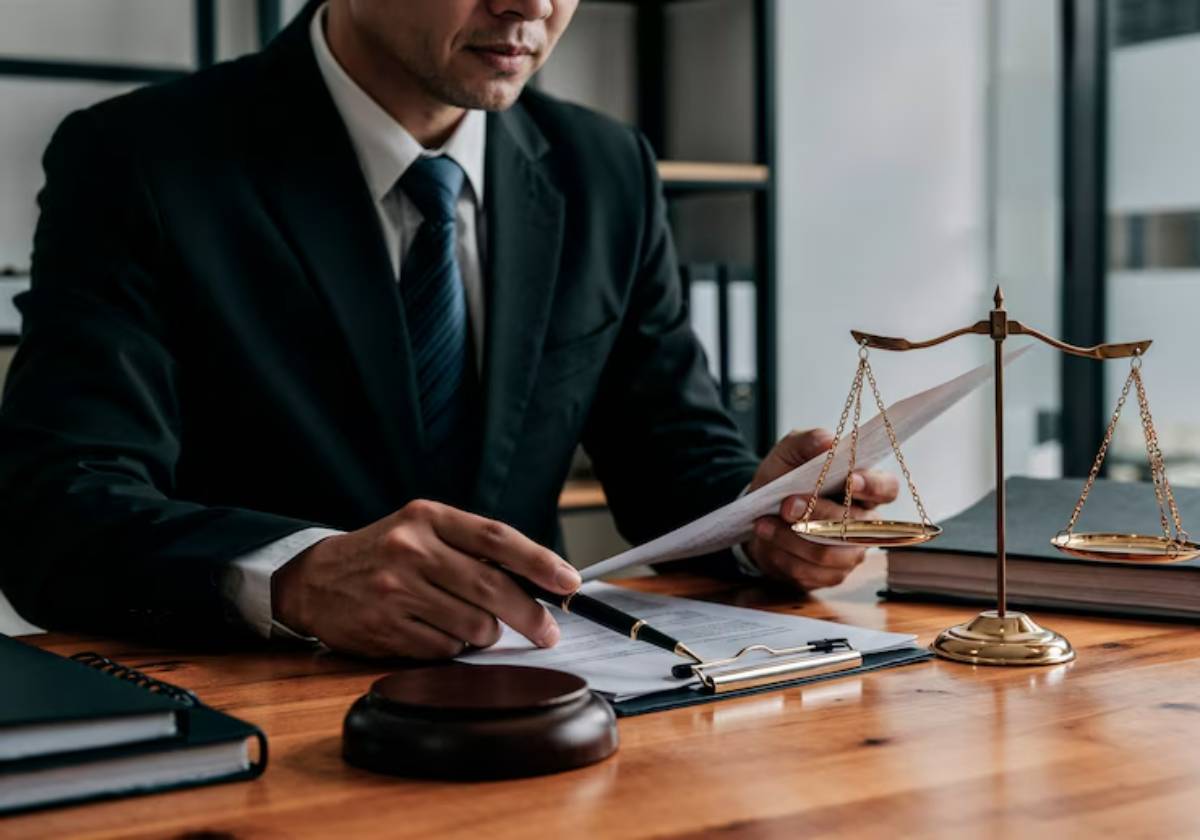 A lawyer in a suit examines documents at a desk, with a gavel and balance scales symbolizing justice in the foreground.