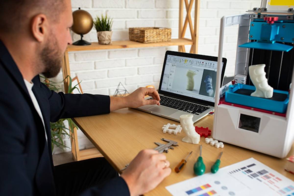 A person sits at a desk, analyzing a 3D model on a laptop. A 3D printer and various tools are arranged on the table.