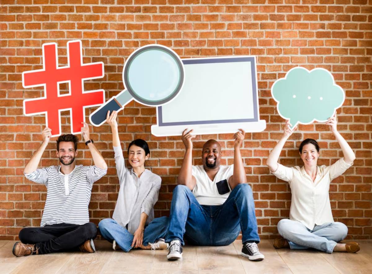 A group of four people sitting on a wooden floor, holding large, playful signs that represent social media concepts against a brick wall.