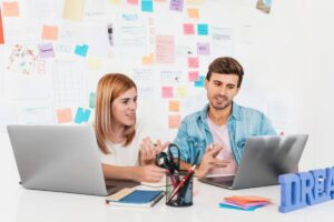 A casual workspace with two people discussing ideas over laptops, surrounded by colorful sticky notes and a DREAM sign.