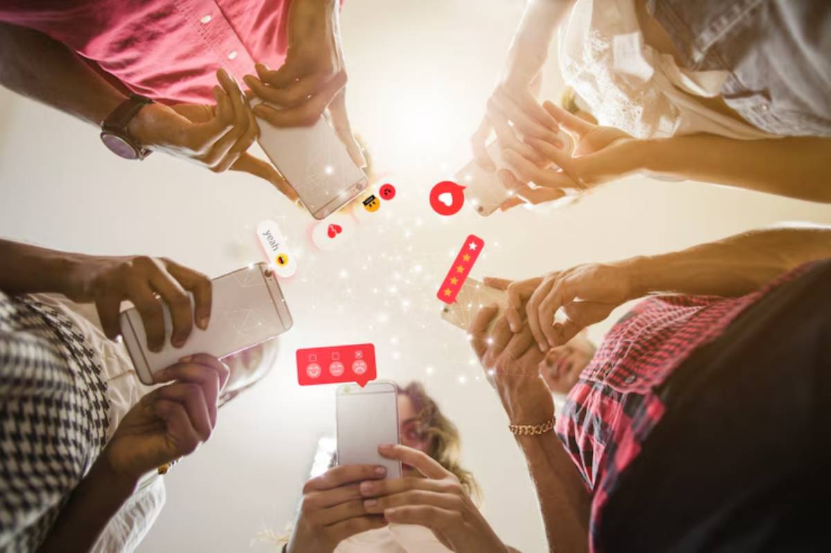 People hold smartphones in a circle, with various social media icons and emojis floating above them, symbolizing digital connection.