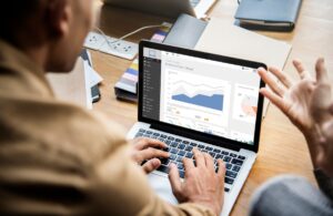 A person uses a laptop displaying various business charts and graphs, while another person gestures during a discussion in a modern workspace.