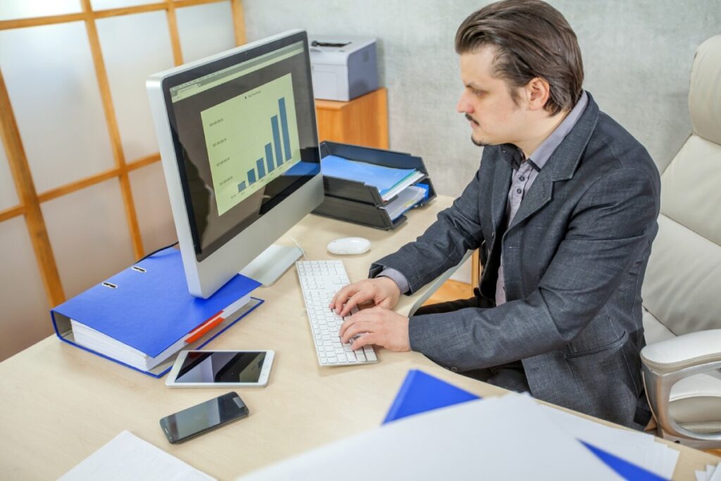 A man in a suit works at a desk with a computer displaying a bar graph, surrounded by folders and smartphone devices.
