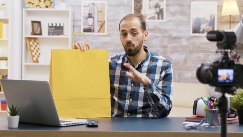 A person in a checkered shirt holds a yellow shopping bag while sitting at a desk with a laptop and a camera focused on them.