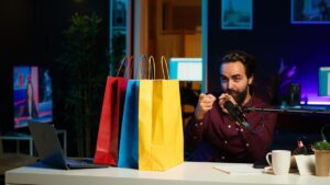 A person points while seated at a desk with three colorful shopping bags, a laptop, and a microphone in a modern studio setting.