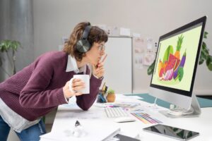 A person with headphones leans over a desk, holding a mug, while intently examining vibrant fruit and vegetable illustrations on a computer screen.