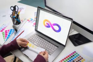 A graphic designer's workspace featuring a laptop displaying an infinity logo, surrounded by color swatches and design tools.