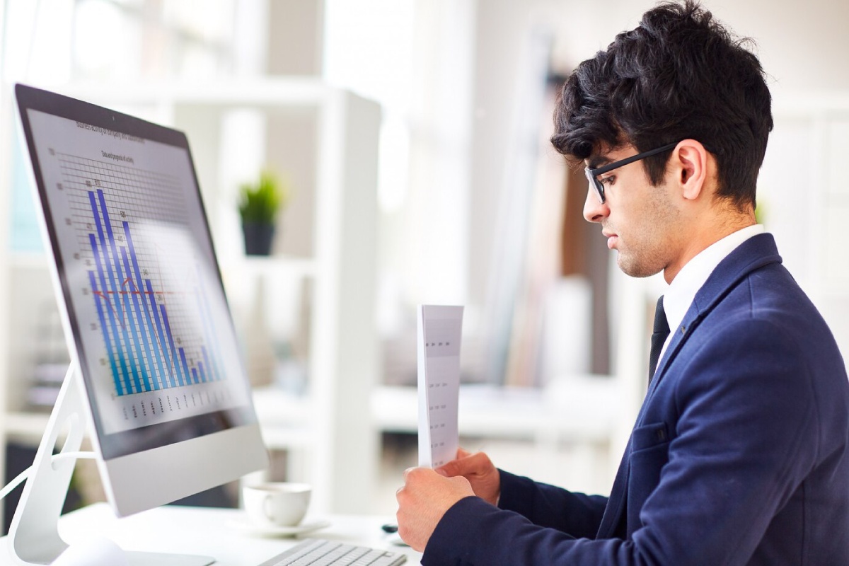 A person in a suit studies a report while seated at a desk, with a computer displaying colorful charts and graphs.