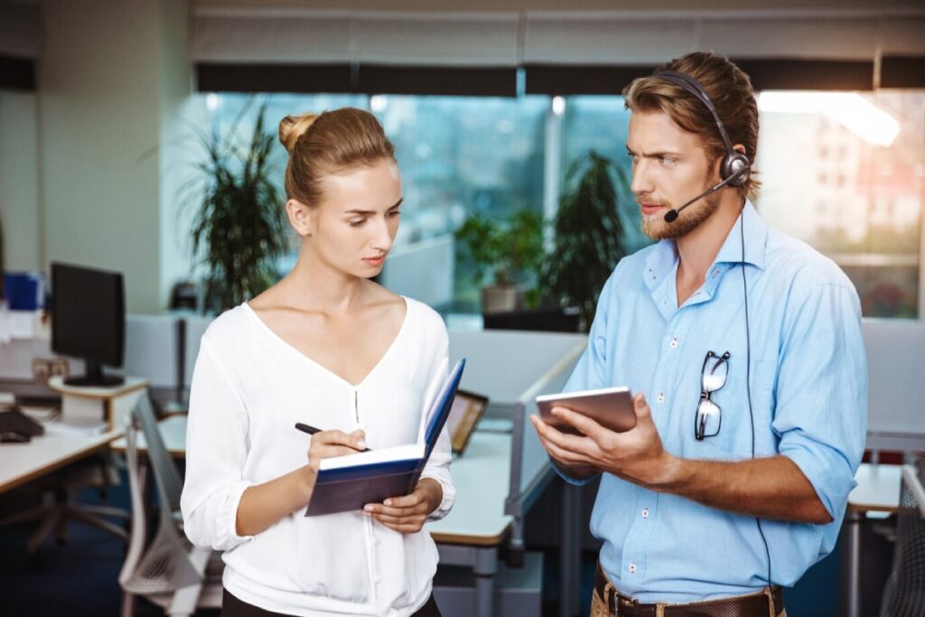 A woman in a white blouse takes notes in a notebook while a man in a blue shirt holds a tablet, engaged in conversation in an office.
