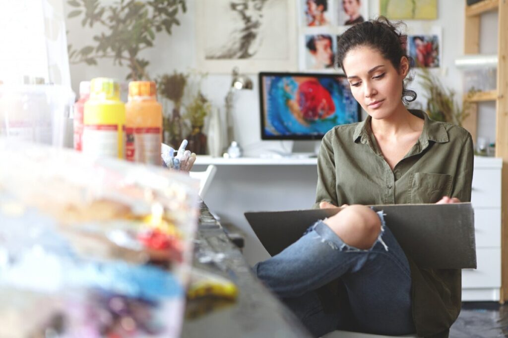 A young artist sits at a cluttered table, holding a sketchpad, surrounded by paint bottles and creative supplies in a bright studio.