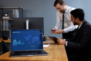 A laptop displays a blue graph highlighting annual statistics and market centers, while two men discuss in a modern office setting.