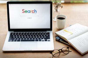 A laptop displaying a search engine page beside a notebook, glasses, and a cup of coffee on a wooden desk.