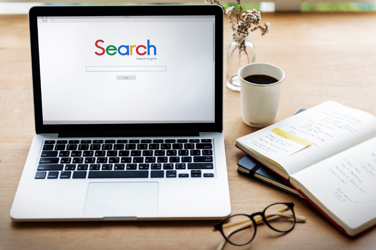 A laptop displaying a search engine page beside a notebook, glasses, and a cup of coffee on a wooden desk.