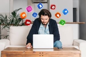 A person sitting on a couch, focused on a laptop, surrounded by colorful social media icons in a modern living space.