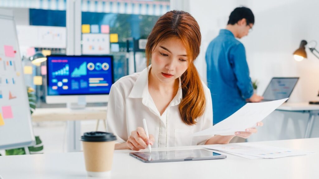 A woman in a white shirt uses a tablet while reviewing papers in a modern office with colorful charts on a screen behind her.