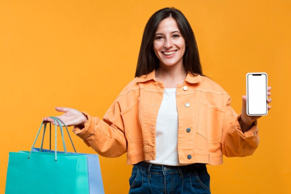 A person wearing an orange jacket holds colorful shopping bags in one hand and a smartphone in the other against a bright yellow background.