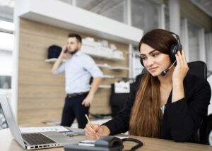 A professional woman wearing a headset takes notes at her desk, while a man stands in the background talking on a phone.