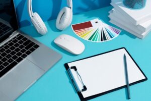 A minimalistic workspace featuring a laptop, headphones, a mouse, color swatches, a clipboard, and a pen on a vibrant turquoise desk.