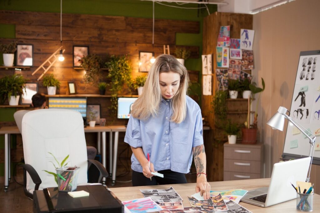 A person in a blue shirt examines printed materials on a desk in a greenery-filled office space, with computers in the background.