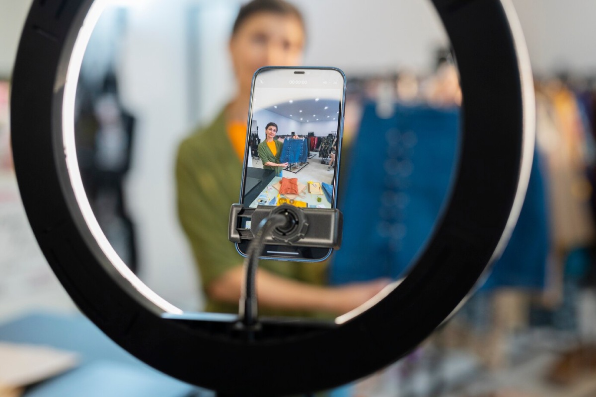 A smartphone on a tripod focused on a table displaying colorful clothing and accessories, framed by a ring light in a shop.