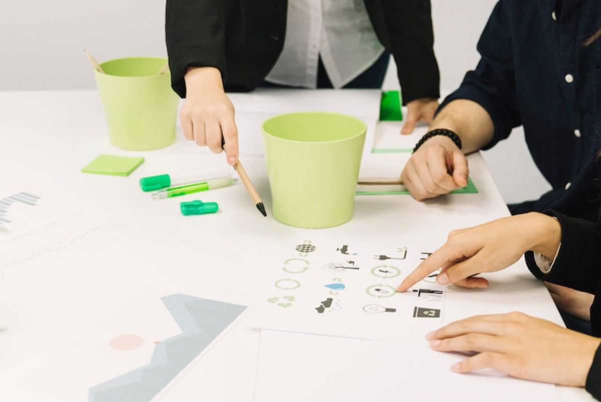 Two people are analyzing graphics on a table, one pointing at eco-friendly icons while surrounded by green stationery and a bucket.