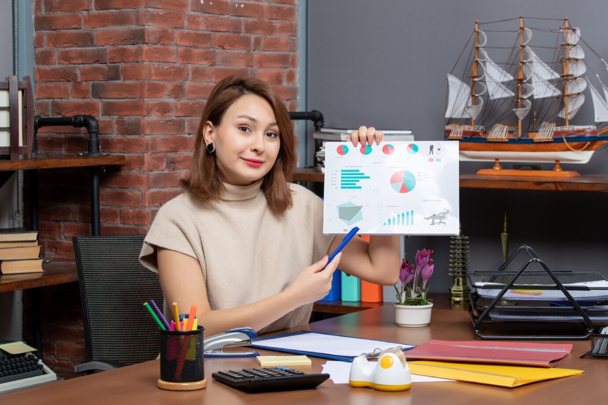 A person sits at a desk, holding a report with charts and graphs, surrounded by office supplies and decor in a brick-walled workspace.