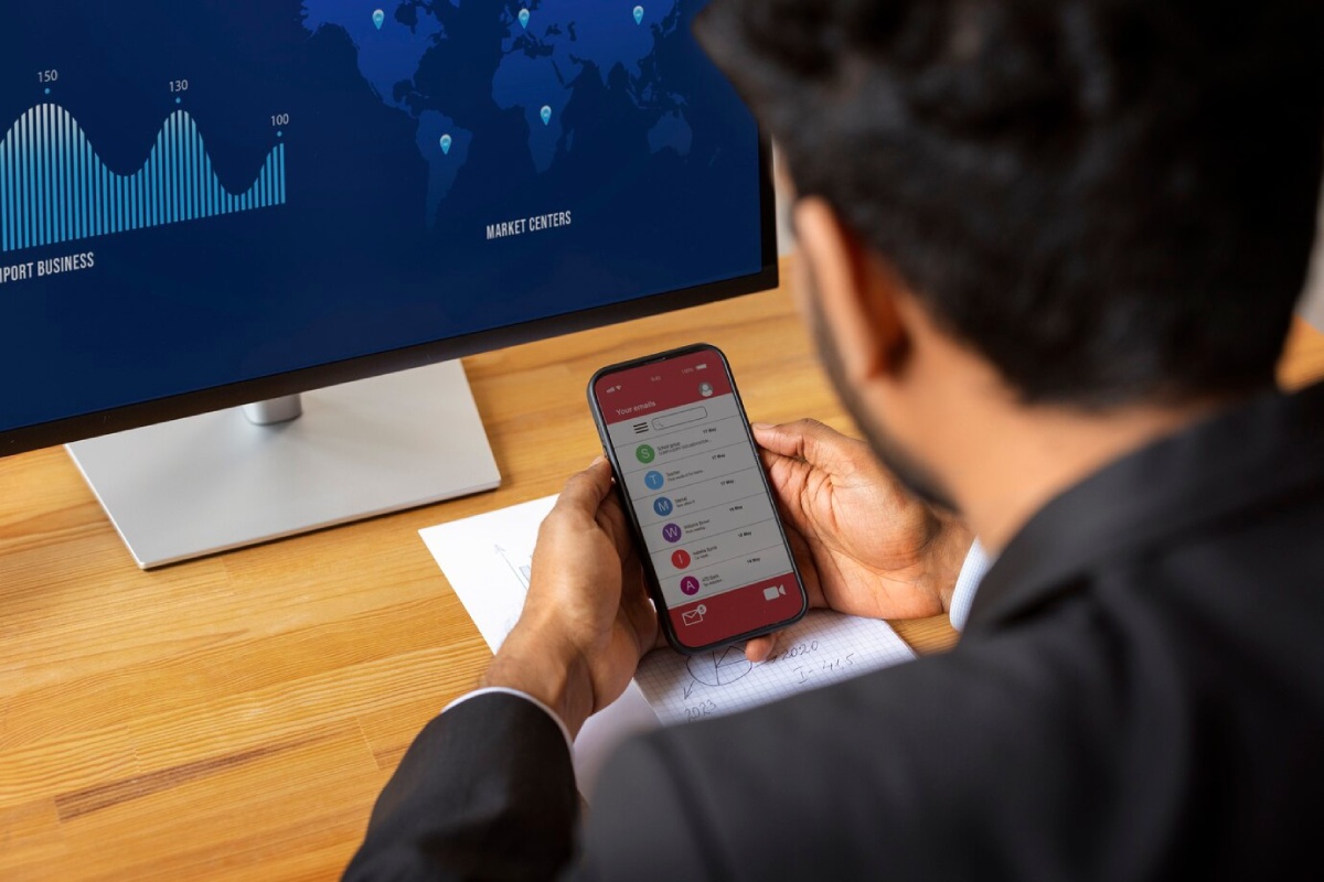 A businessman in a suit checks his emails on a smartphone while analyzing market data displayed on a monitor.