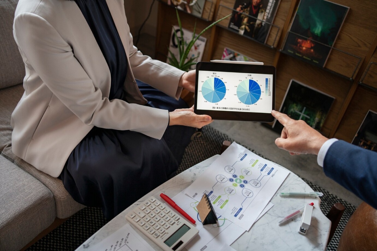 A business meeting where a woman shows pie charts on a tablet, while a man points, surrounded by documents and a calculator.