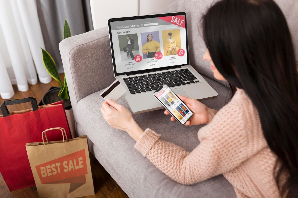 A woman in a cozy sweater shops online with a laptop and smartphone, surrounded by shopping bags. A credit card is in hand.