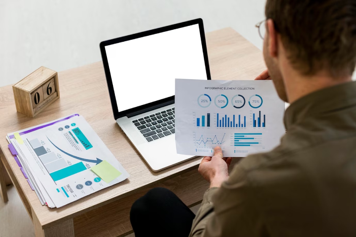 A person reviews an infographic with various charts while sitting at a desk with a laptop and stacked documents.