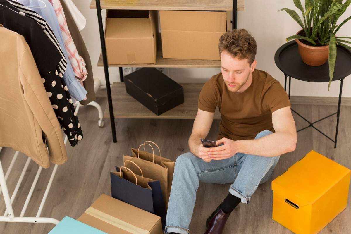 A person sits on the floor surrounded by bags and boxes, focused on their phone, with a clothing rack and potted plant nearby.