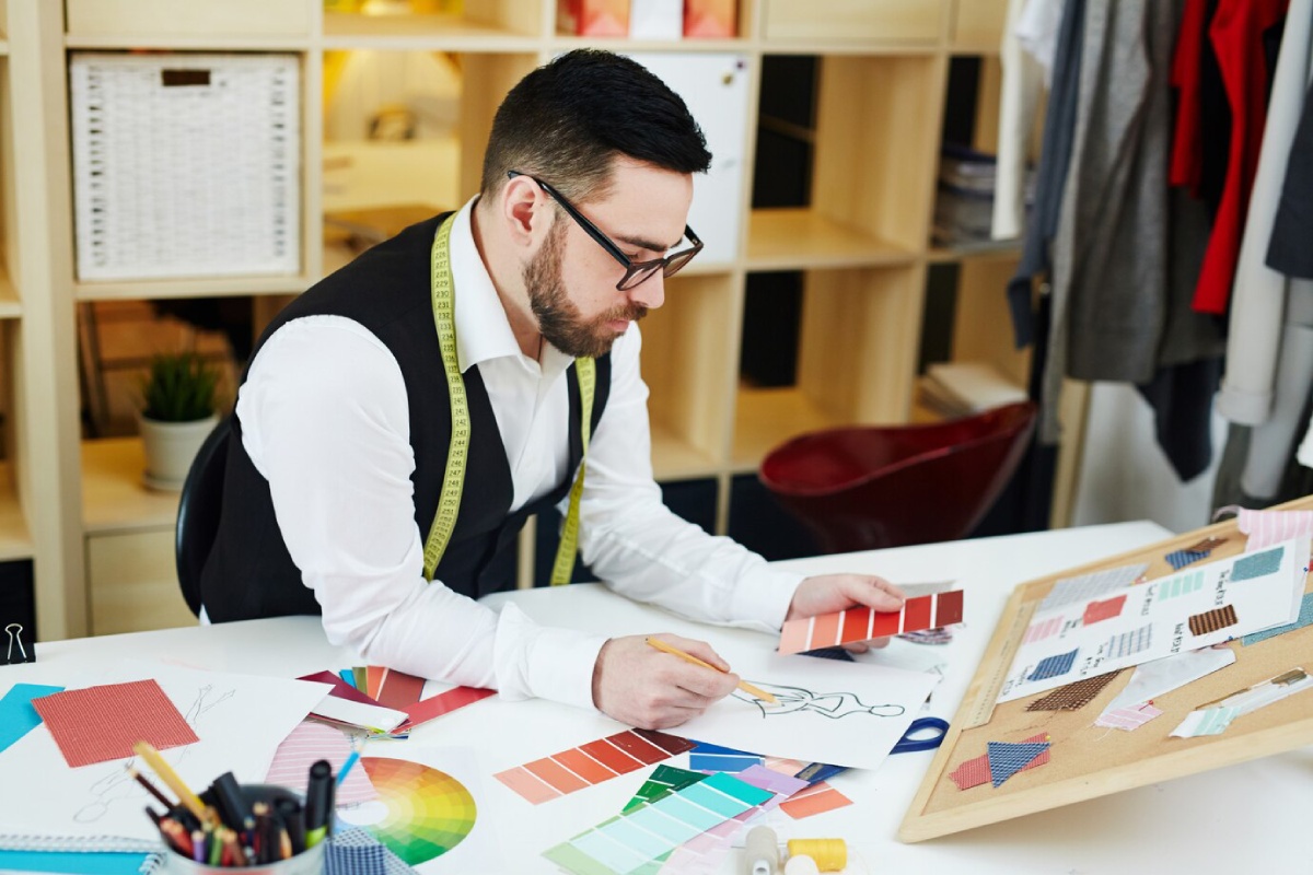 A designer sketching fashion ideas at a table, surrounded by fabric swatches, color palettes, and sketches in a creative workspace.