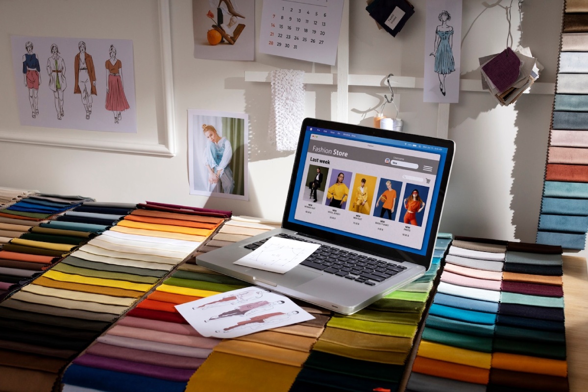 A laptop displaying a fashion website sits on a table covered with vibrant fabric swatches, sketches, and design ideas.