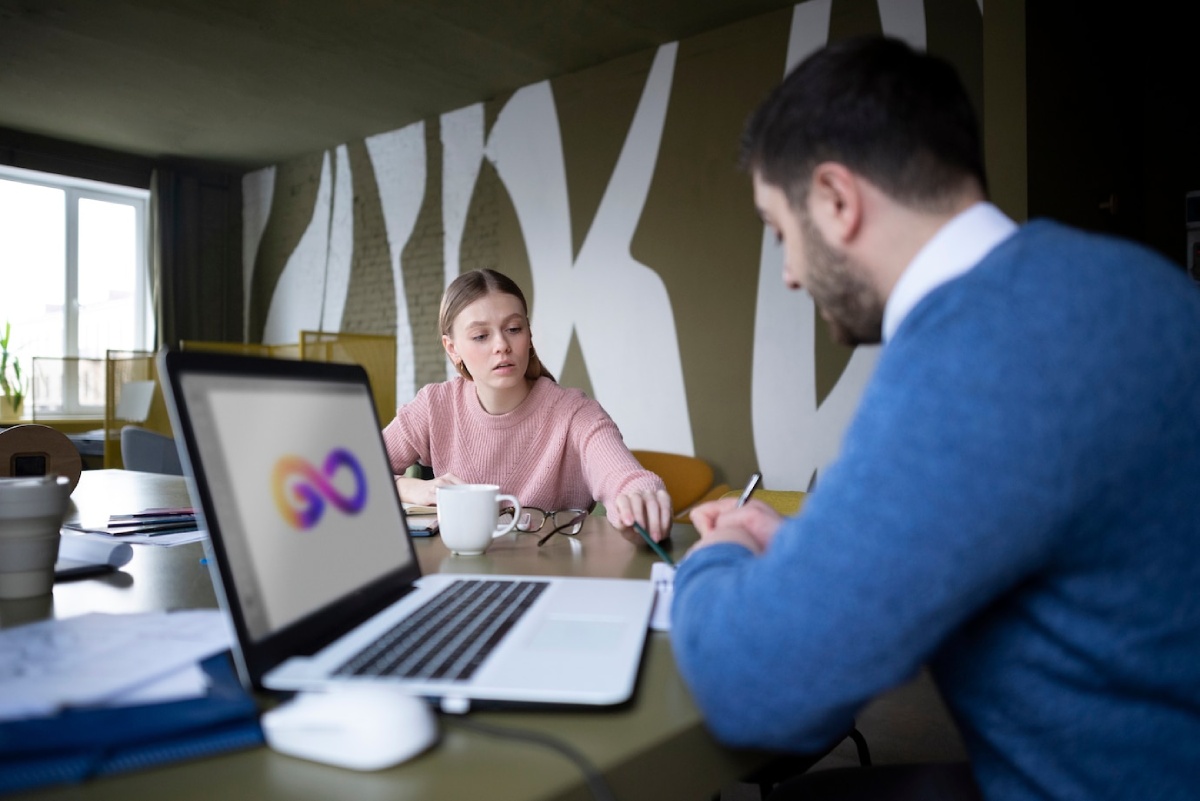 A cozy meeting space with a laptop displaying a colorful logo, two individuals engaged in conversation, and a coffee cup on the table.