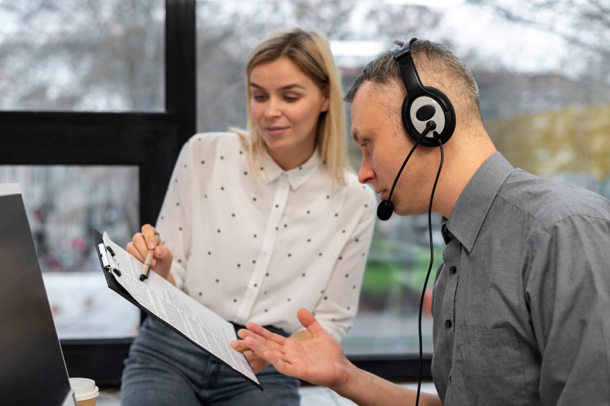 A man wearing headphones engages in a discussion with a woman, who holds a clipboard with papers, in a modern office setting.
