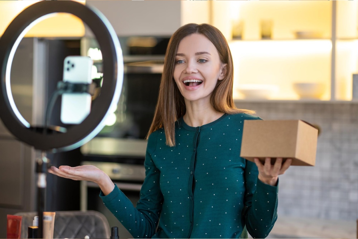A person in a green polka-dot sweater holds a brown package while gesturing, with a ring light and kitchen backdrop.