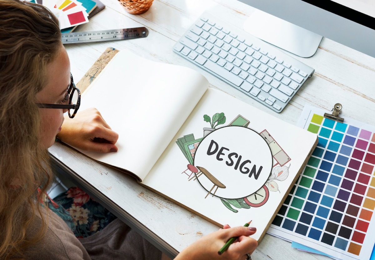 A person is sketching a design concept in a notebook, surrounded by color swatches, a computer, and art supplies on a desk.