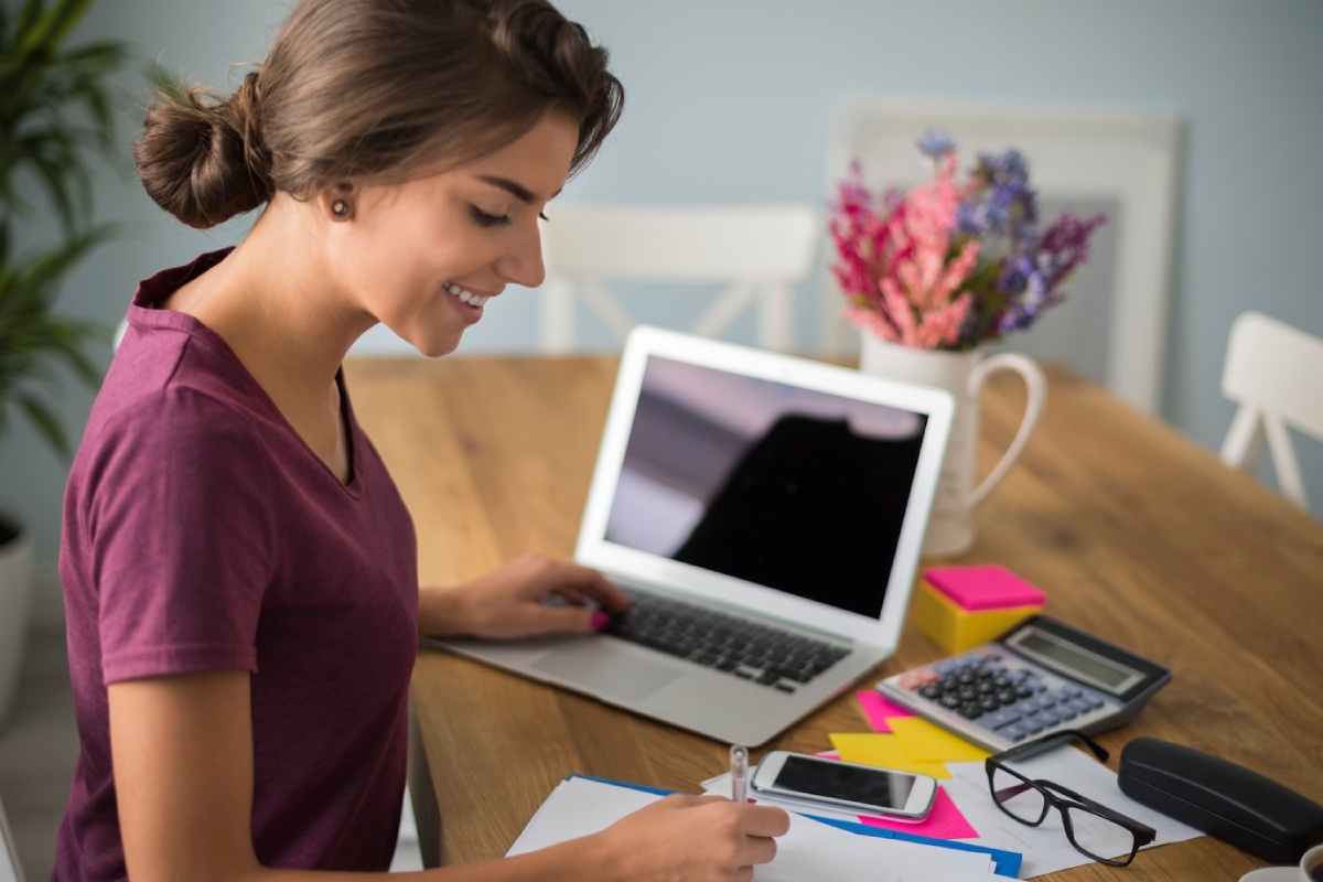 A woman sits at a wooden desk working on a laptop, surrounded by stationery, a calculator, and a decorative flower vase.