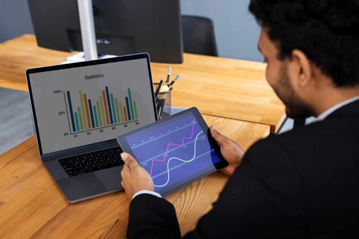 A person in a suit analyzes data on a tablet, while a laptop displays colorful bar graphs labeled Statistics on a wooden desk.