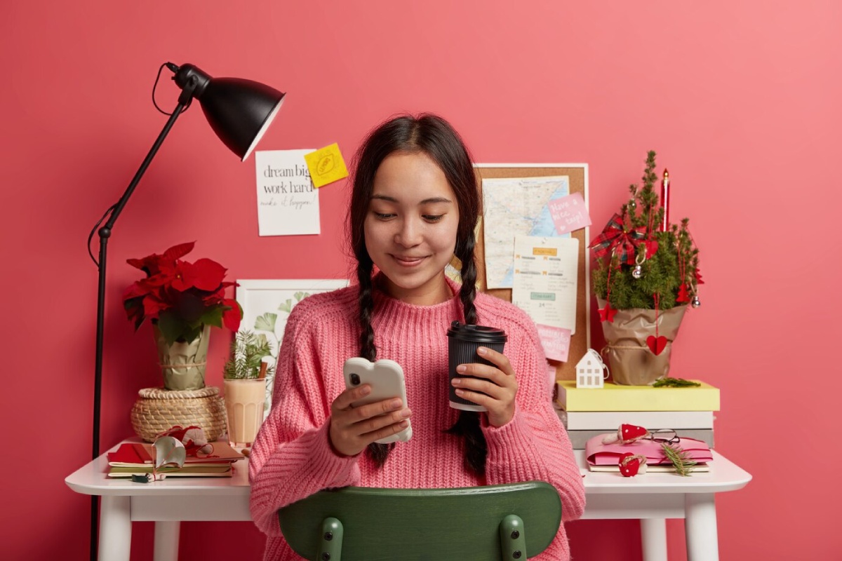 A person in a pink sweater sits at a desk, holding a coffee cup while browsing a smartphone, surrounded by holiday decorations.