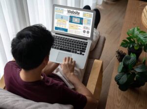 A person sitting on a couch designs a website on a laptop, surrounded by plants in a bright, modern living space.