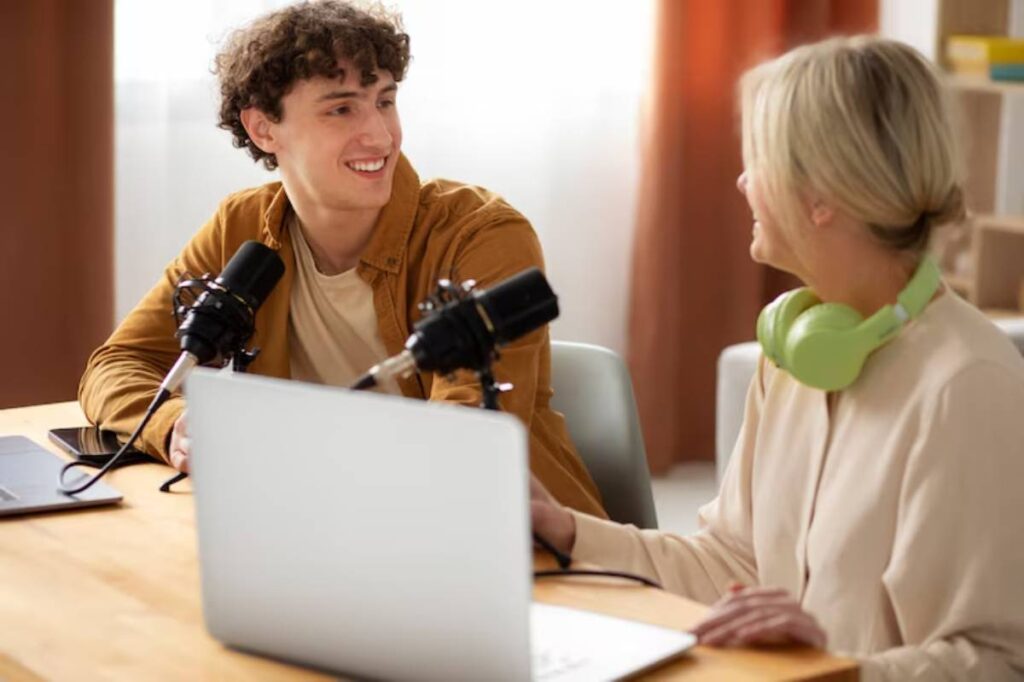 Two individuals sit at a wooden table with microphones and a laptop, engaged in conversation in a bright, modern room.