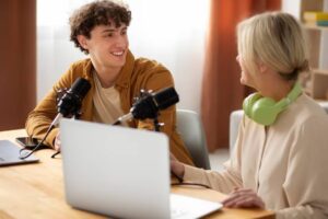 Two individuals sit at a wooden table with microphones and a laptop, engaged in conversation in a bright, modern room.